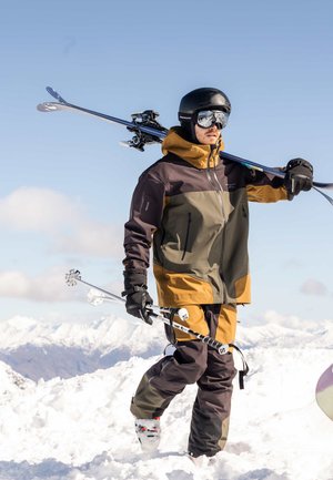 Un skieur portant un casque, des lunettes de ski et une tenue d’hiver transporte ses skis et ses bâtons sur un sommet enneigé.
