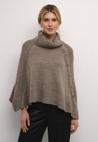 Bež villane ponšo kõrge kraega, millel on ribatakistused ja viis hõbedast nuppu ühe külje peal. Pehme tekstuur, vaba lõige.