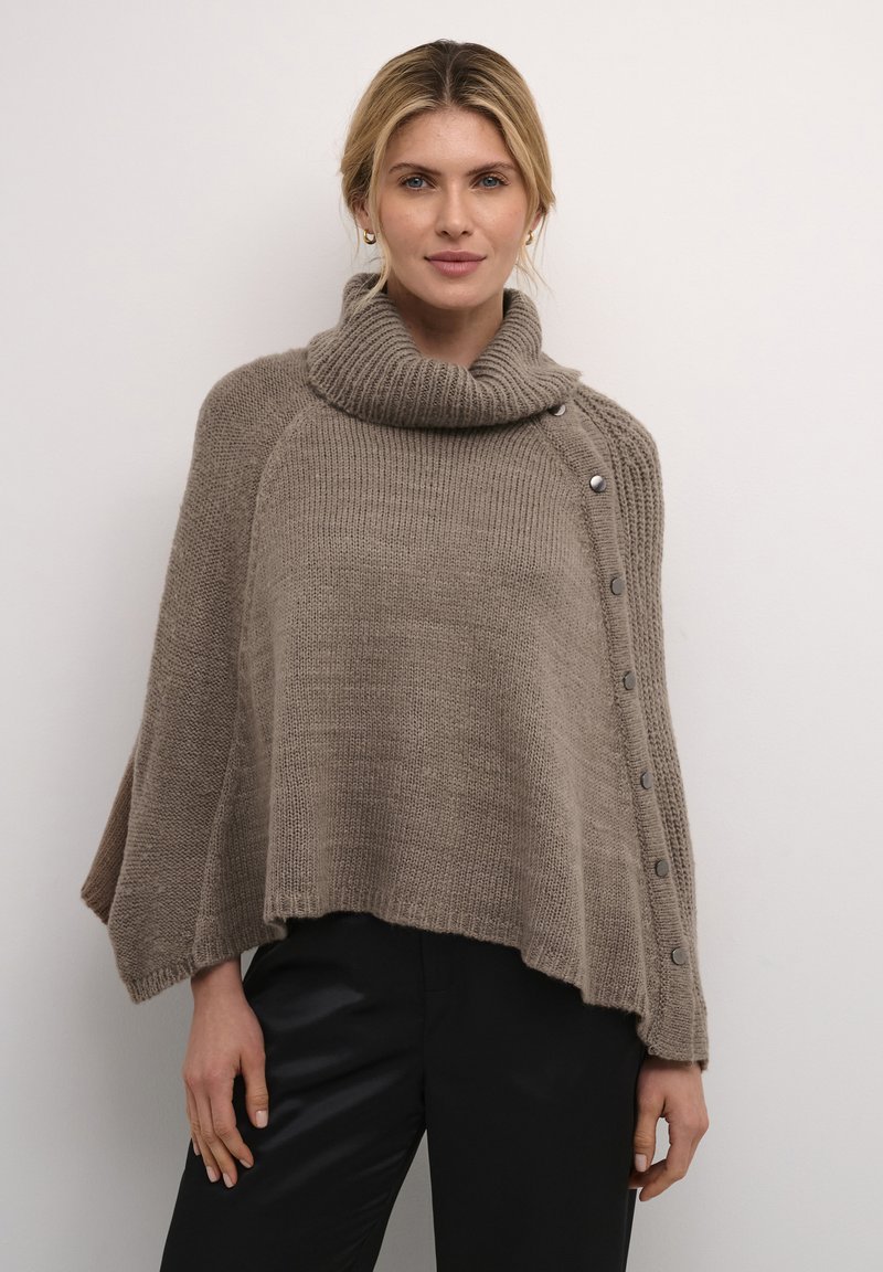 Bež villane ponšo kõrge kraega, millel on ribatakistused ja viis hõbedast nuppu ühe külje peal. Pehme tekstuur, vaba lõige.