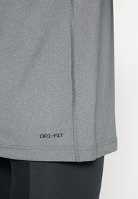 Primer plano del dobladillo de una camiseta deportiva gris con el logo "DRI-FIT" encima de unos pantalones negros, destacando la textura del tejido y los detalles de la costura.