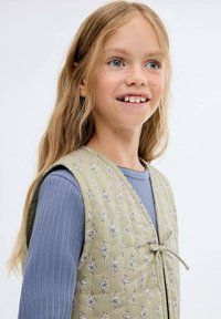 Grön mönstrad väst med knytning över en ljusblå, randig långärmad skjorta. Vesten har en blommig design och en mjuk känsla.