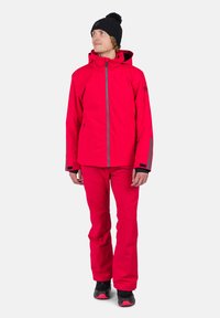Chaqueta impermeable roja con capucha, detalles grises y cremallera frontal, combinada con pantalones rojos a juego. Gorro de lana negro y zapatos.
