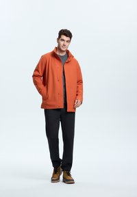Orange waterproof jacket with a high collar, grey shirt underneath, black pants, and brown boots, standing against a white background.