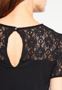 Černý top s krajkovým zadním dílem a designem ve tvaru klíčové dírky, krátkými rukávy a knoflíkovým zapínáním u krku. Měkká textura s propracovanými vzory.