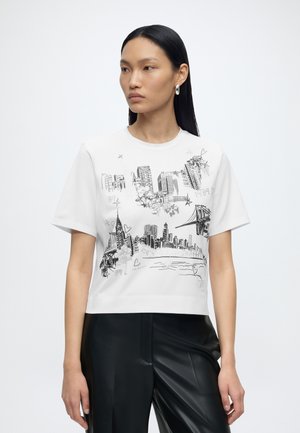 Femme aux longs cheveux foncés portant un t-shirt court blanc avec des croquis noirs d'une silhouette de ville et d'un pont, assorti à un pantalon en cuir noir.