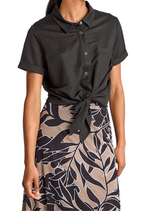 Femme portant une chemise noire à manches courtes boutonnée, nouée à la taille, et une jupe beige avec de grands motifs de feuilles noires.