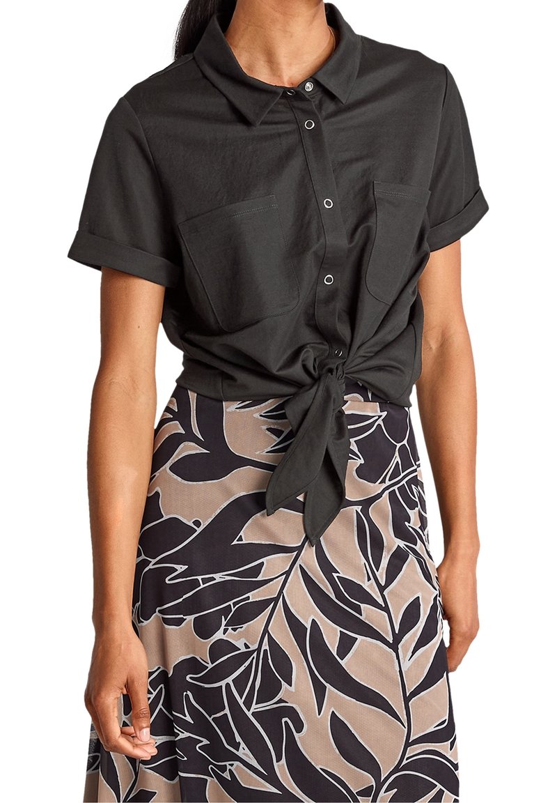 Femme portant une chemise noire à manches courtes boutonnée, nouée à la taille, et une jupe beige avec de grands motifs de feuilles noires.