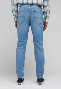 Jean bleu clair avec une coupe slim, design à cinq poches, couture subtile et une étiquette en cuir à la taille, associés à des baskets noires.