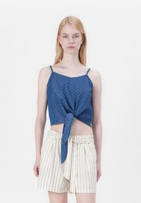 Top scurt din denim albastru cu detaliu legat în față și bretele subțiri, asortat cu șorturi albe cu dungi, având talie cu șnur.