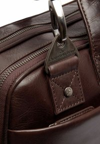 Bruine leren tas met een metalen rits, gestikte accenten en een merklabel met een karabijnhaak. Gladde textuur met een matte afwerking.