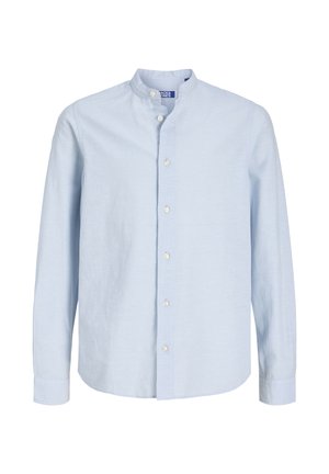 Ragazzo biondo in piedi con una camicia azzurro chiaro abbottonata, pantaloni beige e sneakers beige su uno sfondo chiaro uniforme.