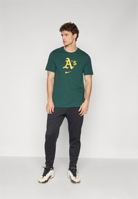 Zelené bavlnené tričko s žltým logom "A's" a emblémom Nike, spárované s čiernymi teplákmi a bielymi teniskami. Model stojí na neutrálnom pozadí.