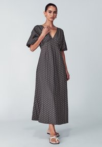 Szara maxi sukienka w kropki z dekoltem w kształcie litery V i krótkimi bufiastymi rękawami, wykonana z lekkiego materiału. Noszona z białymi sandałami.