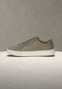 Baskets basses en cuir beige avec semelle blanche et lacets, reposant sur une plateforme ronde claire sur un fond beige uni.