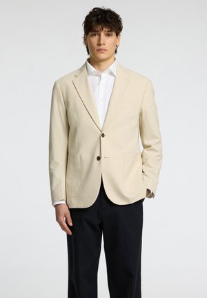 Béžový pruhovaný blazer s klopovými límci, dvojitým zapínáním na knoflíky a dvěma předními kapsami, kombinovaný s bílou košilí a tmavými kalhotami.