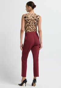 Woman with hair in bun wears sleeveless leopard print top, burgundy slim-fit pants, and black high heels, standing against plain background.
