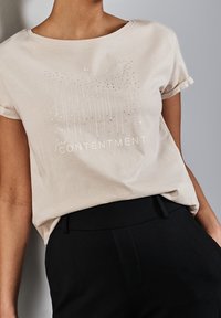 Beige bomullströja med korta ärmar, med en grafisk design av vertikala linjer och texten "find CONTENTMENT" i silvertryck.