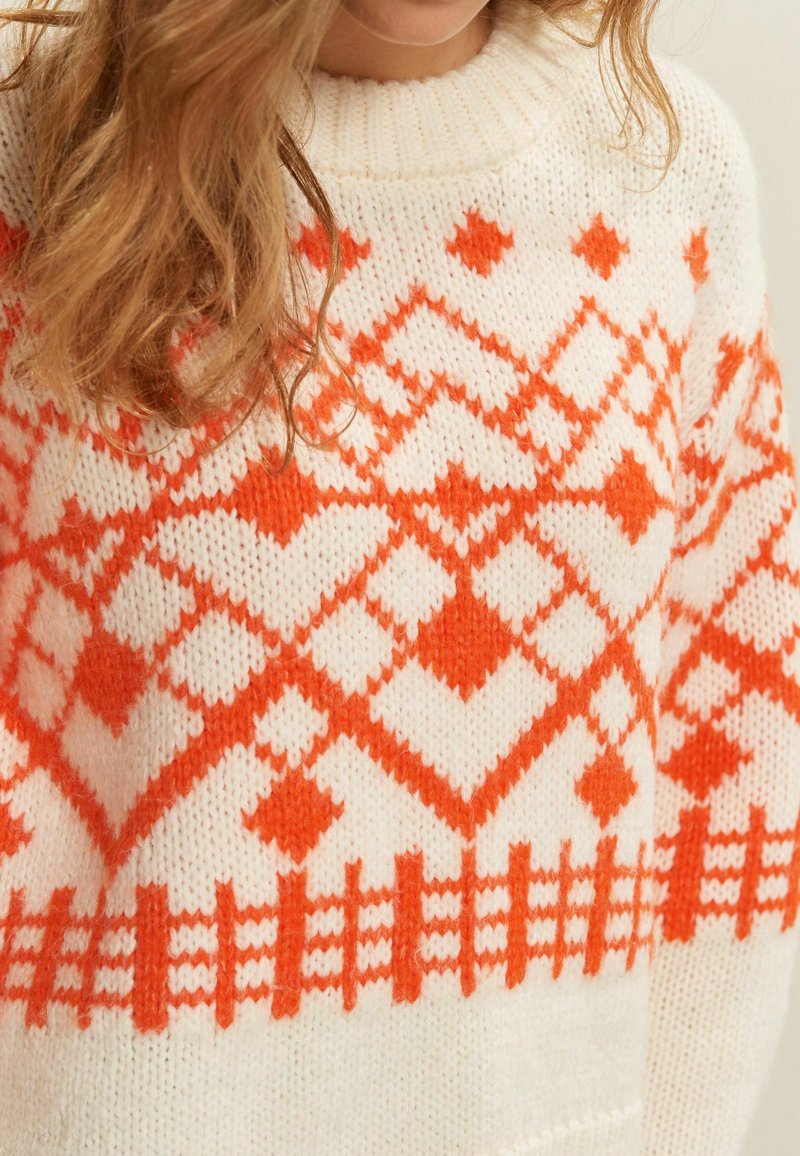 Strickpullover mit cremefarbener Grundfarbe und kräftigen orangen geometrischen Mustern, darunter Herzen und Rauten, sowie einem strukturierten, gerippten Kragen.