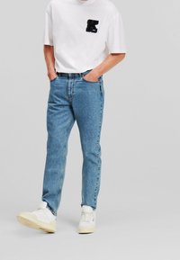 Jeans de mezclilla de color azul claro con corte recto, combinados con una camiseta blanca que presenta una "K" negra bordada en el pecho. Zapatillas blancas.