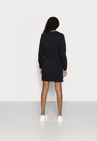 Svart sweatshirtklänning med långa ärmar, figurnära midja och en mjuk textur. Stylad med vita sneakers mot en neutral bakgrund.