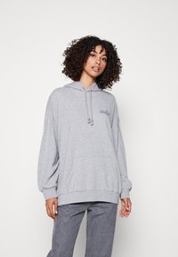 Sudadera gris claro con un bolsillo frontal, capucha con cordón, corte holgado y un logotipo azul bordado en el pecho, combinada con jeans gris de pierna ancha.