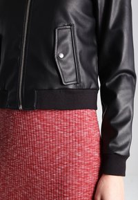 Černá kožená bomber bunda se zapínáním na zip vpředu, žebrovanými manžetami a náprsní kapsou, v kombinaci s červenou žebrovanou sukní s texturou.