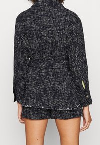 Veste texturée bleu marine avec une taille ceinturée, dotée de deux accents de poignets boutonnés et de détails d'ourlet effiloché.