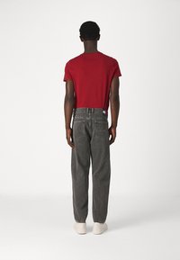 Hombre de pie, de espaldas, vistiendo una camiseta roja, jeans negros desgastados con parche de marca y zapatillas blancas, sobre un fondo blanco liso.