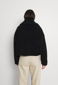 Czarna krótka kurtka z polaru o fakturowanym wykończeniu, z dużym kołnierzem i długimi rękawami, w połączeniu z jasnymi, prostymi spodniami.