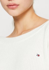 Biały sweter z dzianiny o szerokim dekolcie, z małym, wielokolorowym logo na lewej piersi. Gładka tekstura i luźny krój.