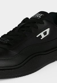 Baskets Diesel noires avec semelle épaisse, lacets noirs, logo blanc sur le côté et marque Diesel sur la languette sur un fond uni.