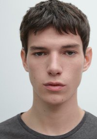 Portret osoby o krótkich, potarganych ciemnych włosach i neutralnym wyrazie twarzy, ubranej w zwykły ciemnoszary t-shirt na jasnym tle.