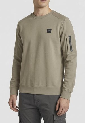 Uomo che indossa una felpa beige a manica lunga con una piccola toppa nera "AM" sul petto e una tasca con zip sulla manica sinistra.