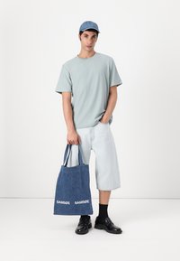 Light blue cotton t-shirt, light denim shorts, dark shoes. Carrying a blue denim tote with white "SAMSØE" logo. Blue cap. Minimalist design.