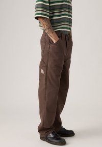 Pantalons cargo marron avec poches latérales, fabriqués en coton. Caractérisés par une coupe décontractée et une étiquette de marque visible. Portés avec des chaussures noires.