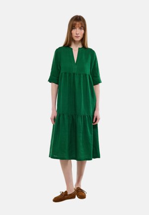 Femme debout portant une robe verte à volants longueur genou avec des manches longueur coude et des mocassins marron à lacets.