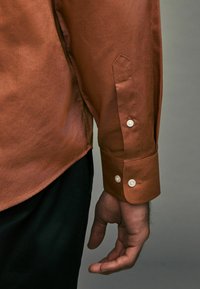 Braunes langärmeliges Hemd mit glatter Textur, ausgestattet mit einem geknöpften Manschettenverschluss und einem dezenten Glanz, kombiniert mit schwarzer Hose.