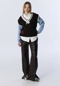 Zwarte gebreide vest met witte, groene en blauwe details, gedragen over een wit shirt; gecombineerd met bruine broek met zwarte leren panelen.