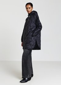 Cappotto imbottito nero trapuntato con cappuccio, dotato di zip frontale, tasche laterali, polsini a coste e vestibilità comoda. Indossato sopra jeans a gamba ampia.