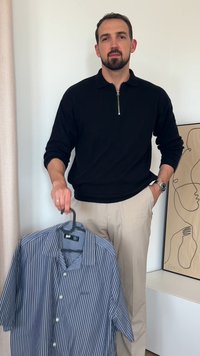 Črna zip-up jopica, bež hlače, drži modro in belo črtasto kratkosleečno majico na obešalniku. Minimalistična umetnost v ozadju.
