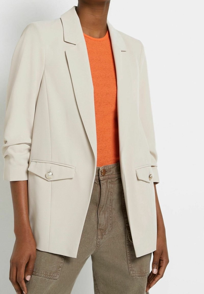Personne portant un blazer crème avec poches à rabat boutonnées, chemise orange et pantalon marron, vue des épaules aux hanches.