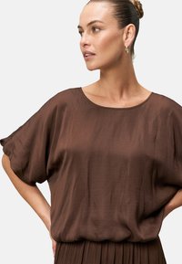 Blusa marrone, morbida e ampia, con maniche larghe e scollatura rotonda. Realizzata in un tessuto leggero e liscio con una leggera lucentezza.