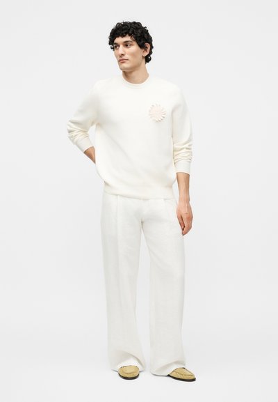 Νέος άνδρας με σγουρά μαλλιά που φορά ολόλευκη εμφάνιση: πουλόβερ με λουλουδάτη λεπτομέρεια, παντελόνι με φαρδιά μπατζάκια και μπεζ παπούτσια slip-on, στέκεται με το χέρι στη μέση.