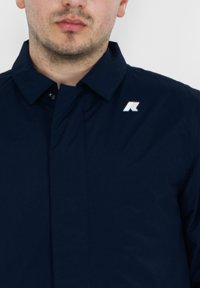Camicia blu navy con colletto strutturato, chiusura a bottoni e un piccolo logo riflettente sul lato sinistro. Texture del tessuto liscia e opaca.