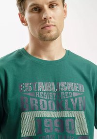 Zielona bawełniana koszulka z wyraźnym grafiką tekstu w szaro-czarnych kolorach, z okrągłym dekoltem i krótkimi rękawami, prezentująca vintage'owy design.