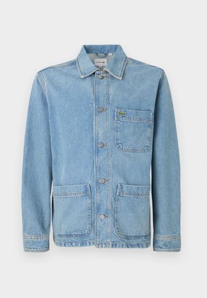 Chaqueta vaquera azul claro con cierre de botones, tres bolsillos delanteros y un pequeño logo de cocodrilo verde en el bolsillo izquierdo del pecho.