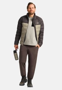 Veste en duvet avec des panneaux noirs et gris clair, t-shirt gris, pantalon marron, portant une bouteille d'eau grise, et portant des bottes noires robustes.