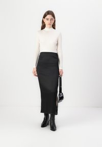 Sydd svart sateng nederdel med sideslitz, parret med en hvid langærmet turtleneck top. Modellen holder en lille sort håndtaske.