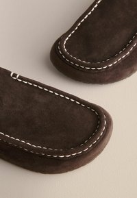 Mocassins en daim marron foncé avec surpiqûres blanches contrastantes le long de l\'orte arrondie et des bords, présentés sur une surface claire.