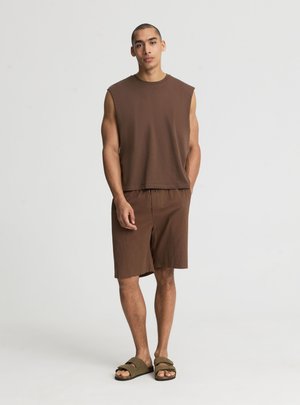 Giovane uomo in piedi che indossa una canottiera marrone senza maniche, pantaloncini marroni abbinati e sandali verde oliva su uno sfondo chiaro monotono.